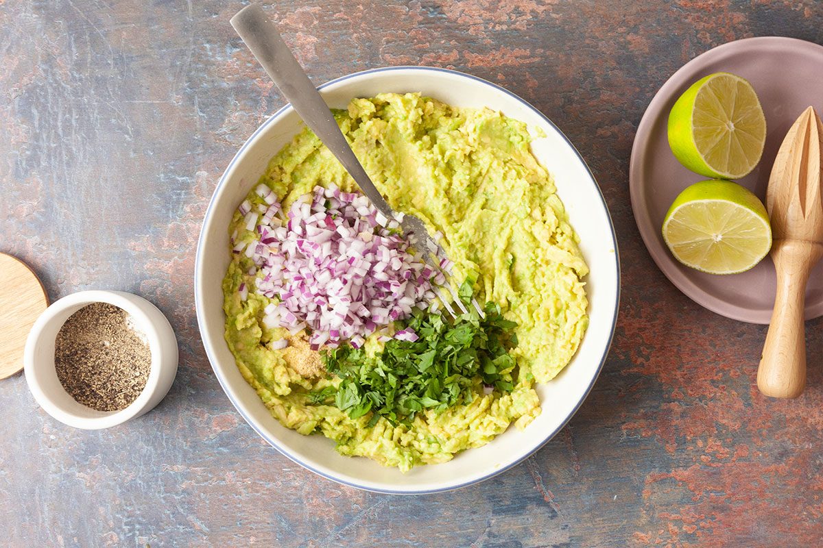 A bowl of mashed avocado topped with chopped red onion and cilantro, with a spoon inside. Next to the bowl are a halved lime with a wooden juicer, and a small bowl of ground pepper.