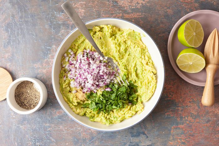 A bowl of mashed avocado topped with chopped red onion and cilantro, with a spoon inside. Next to the bowl are a halved lime with a wooden juicer, and a small bowl of ground pepper.