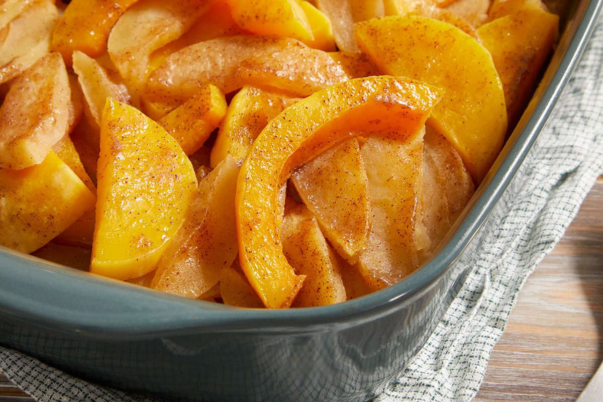 Close-up of sliced apples and squash in a blue baking dish, sprinkled with cinnamon and ready to be baked, sitting on a checkered cloth on a wooden surface.