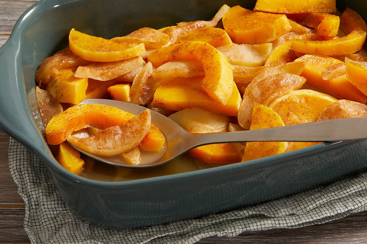 A baking dish filled with sliced yellow squash and onions, seasoned and baked, with a metal serving spoon resting inside. The dish sits on a textured cloth on a wooden surface.