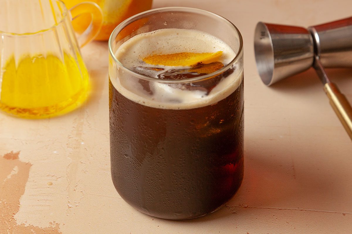 3/4th view angle shot of Carajillo filled in a glass; a small glass pitcher containing liquor and a jigger are visible nearby; all set on a painted surface;