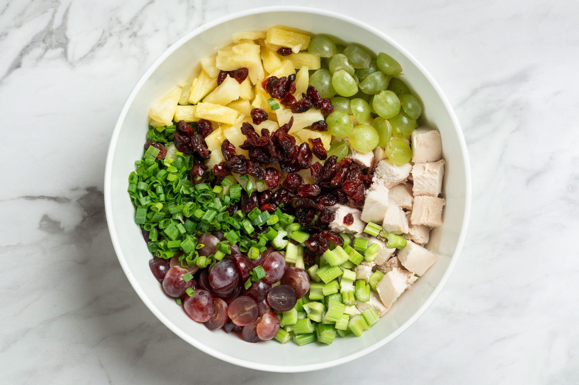 Overhead shot of a large bowl combine the chicken, pineapple, celery, onions, grapes and cranberries; on a marble surface
