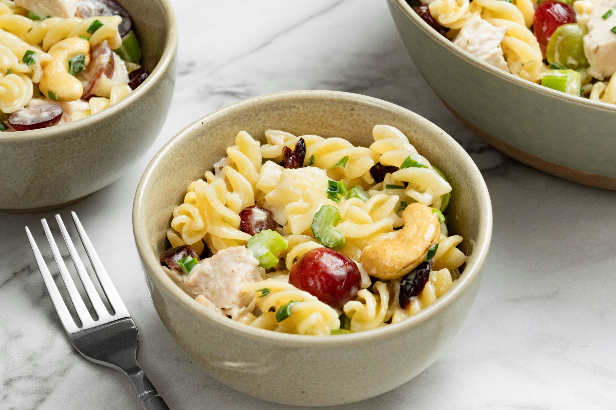 Top view shot of Cashew-Chicken Rotini Salad in a large bowl; served in two small bowls and a fork nearby; all set on a marble surface