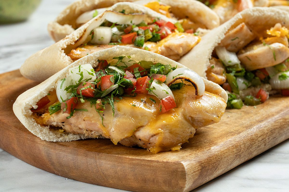 Pita bread filled with grilled chicken, melted cheese, diced tomatoes, onions, and fresh herbs, served on a wooden cutting board.