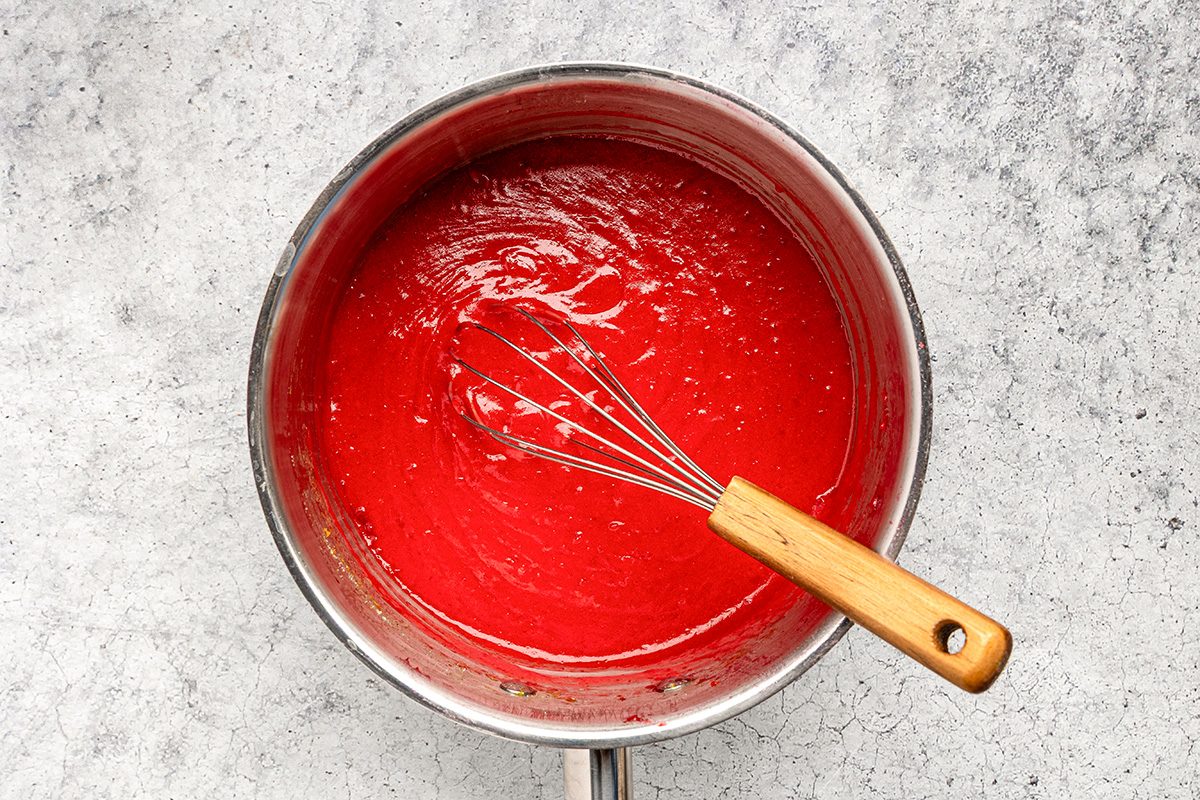 A metal saucepan filled with bright red sauce being whisked with a metal whisk that has a wooden handle, set on a light gray textured surface.