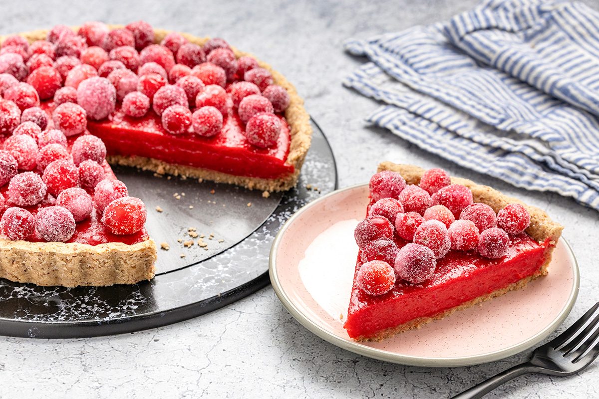 A tart with a crumbly crust and bright red filling topped with sugared cranberries sits on a tray. One slice is served on a pink plate next to a stack of striped napkins.