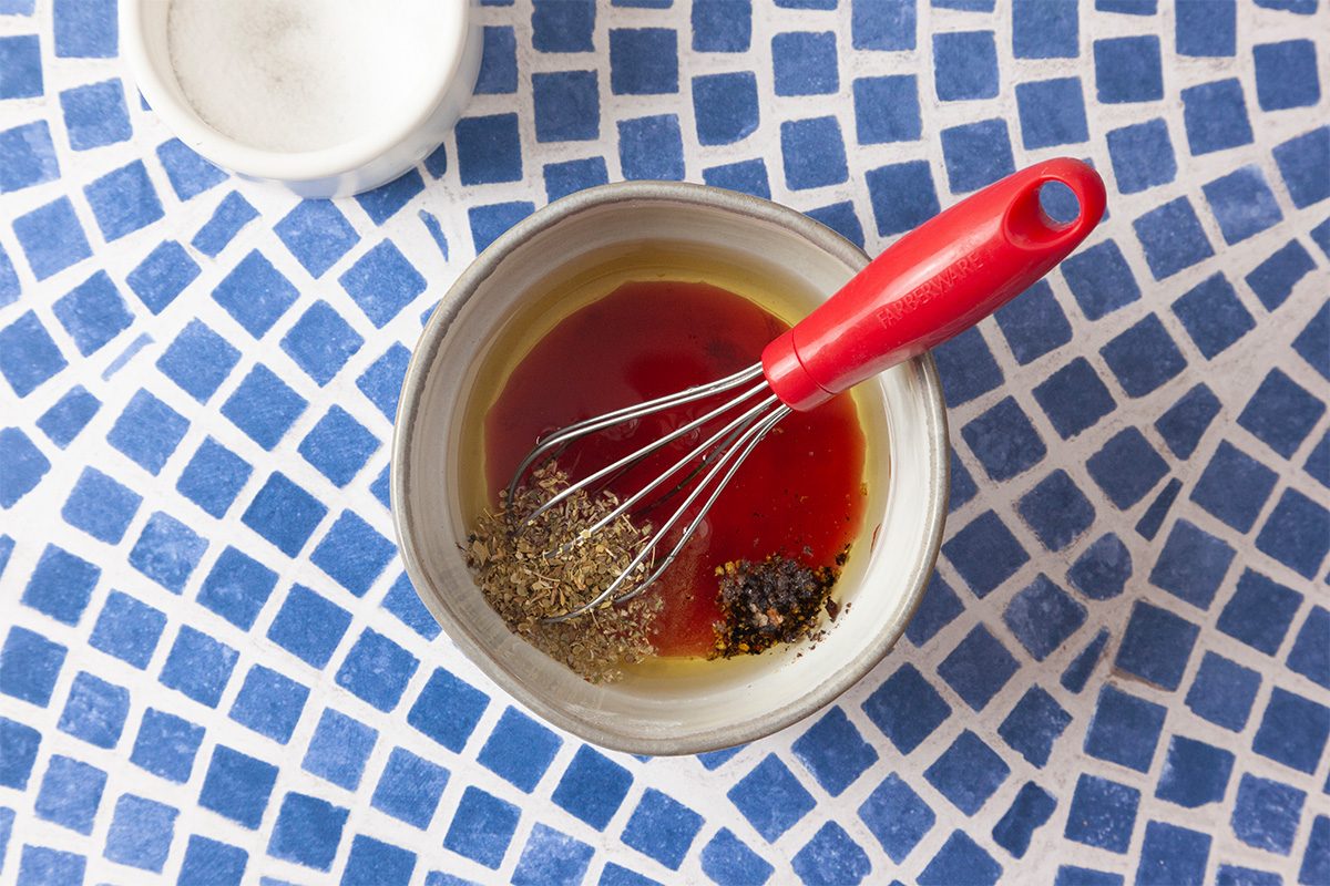 A bowl with oil, vinegar, and seasonings being whisked with a red-handled whisk sits on a blue and white mosaic-tiled surface; a small white bowl is nearby.