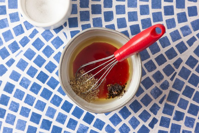 A bowl with oil, vinegar, and seasonings being whisked with a red-handled whisk sits on a blue and white mosaic-tiled surface; a small white bowl is nearby.