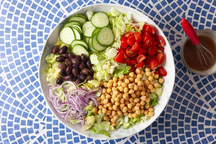 A bowl of salad with sliced cucumbers, cherry tomatoes, chickpeas, black olives, red onion, and lettuce on a blue mosaic table; a small bowl of dressing with a whisk is beside it.