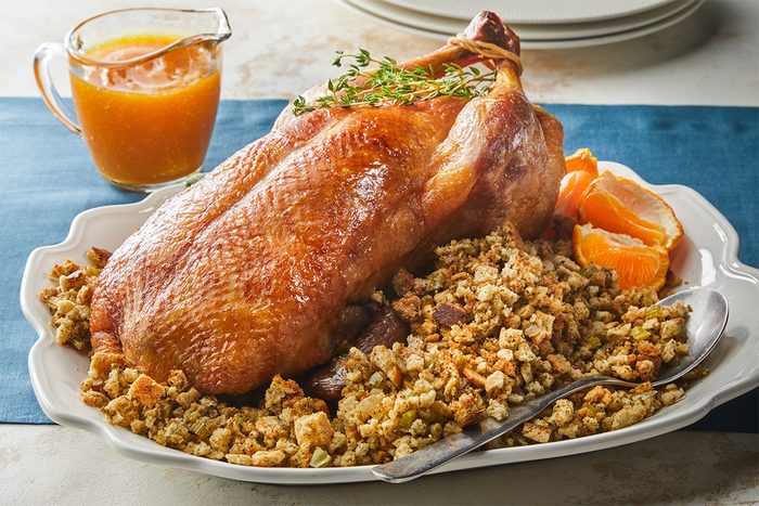 A roasted duck sits on a platter atop a bed of stuffing, garnished with fresh herbs and orange slices. A spoon rests on the side, and a small glass pitcher of sauce is nearby. Plates are stacked in the background.