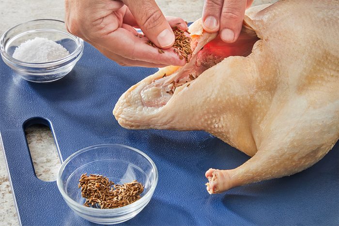 A person stuffs spices into the cavity of a raw whole chicken on a blue cutting board. Two small bowls, one with salt and one with seeds, are nearby.