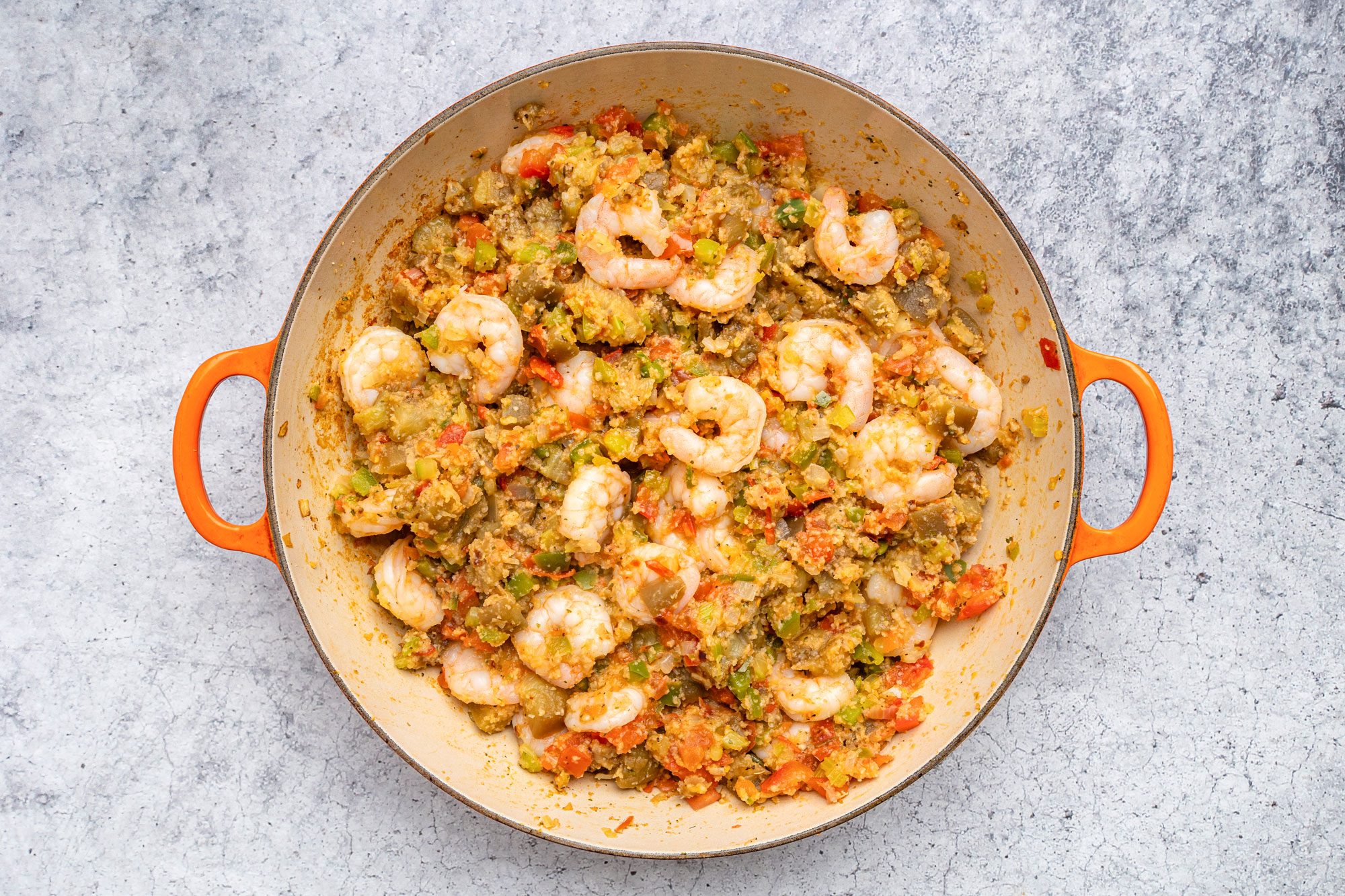 overhead shot of a round orange handled pan filled with shrimp, chopped vegetables, and rice in a saucy mixture, on a gray textured surface