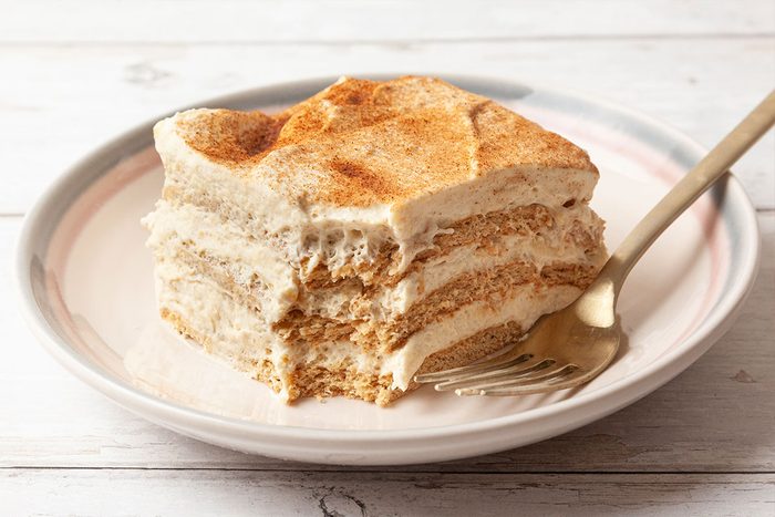 A slice of tiramisu with creamy layers and a dusting of cocoa powder on top, served on a white plate with a fork on a light wooden surface.