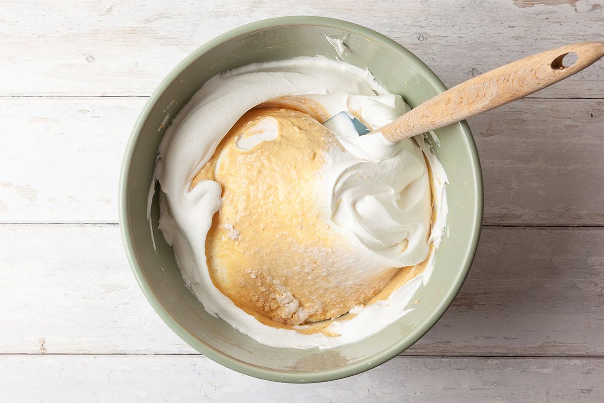 A mixing bowl containing white whipped cream and a yellowish batter, partially combined, with a spatula resting inside. The bowl sits on a light wooden surface.