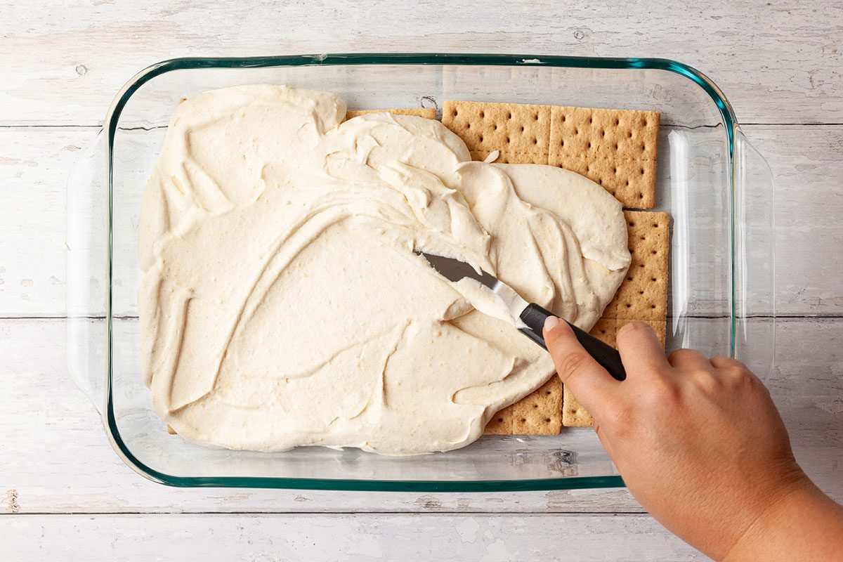 A hand uses an offset spatula to spread creamy filling over a layer of rectangular graham crackers in a glass baking dish on a light wooden surface.