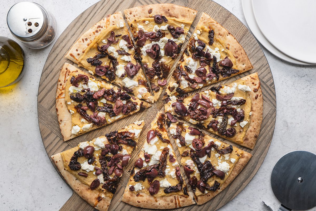 A sliced pizza topped with cheese, caramelized onions, and olives on a round wooden board, with a bottle of olive oil, a pepper shaker, and empty plates nearby.