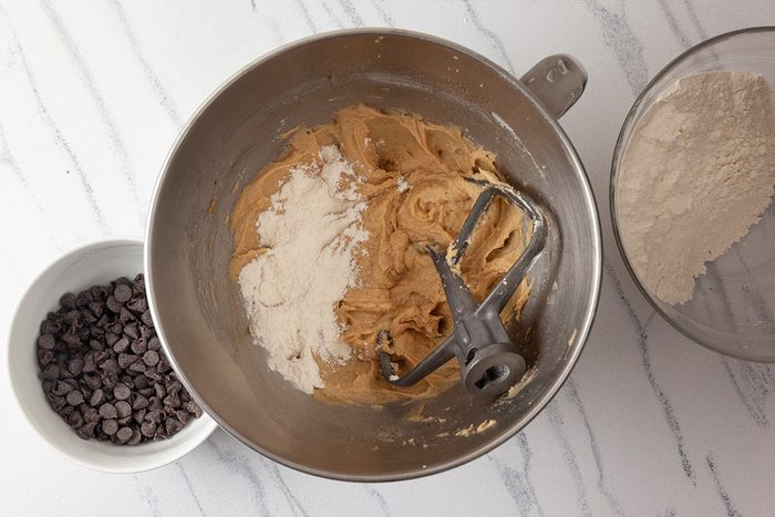A mixing bowl with cookie dough, a beater attachment, and some flour on top. Nearby are a bowl of chocolate chips and a separate bowl of flour, all on a white marble countertop.