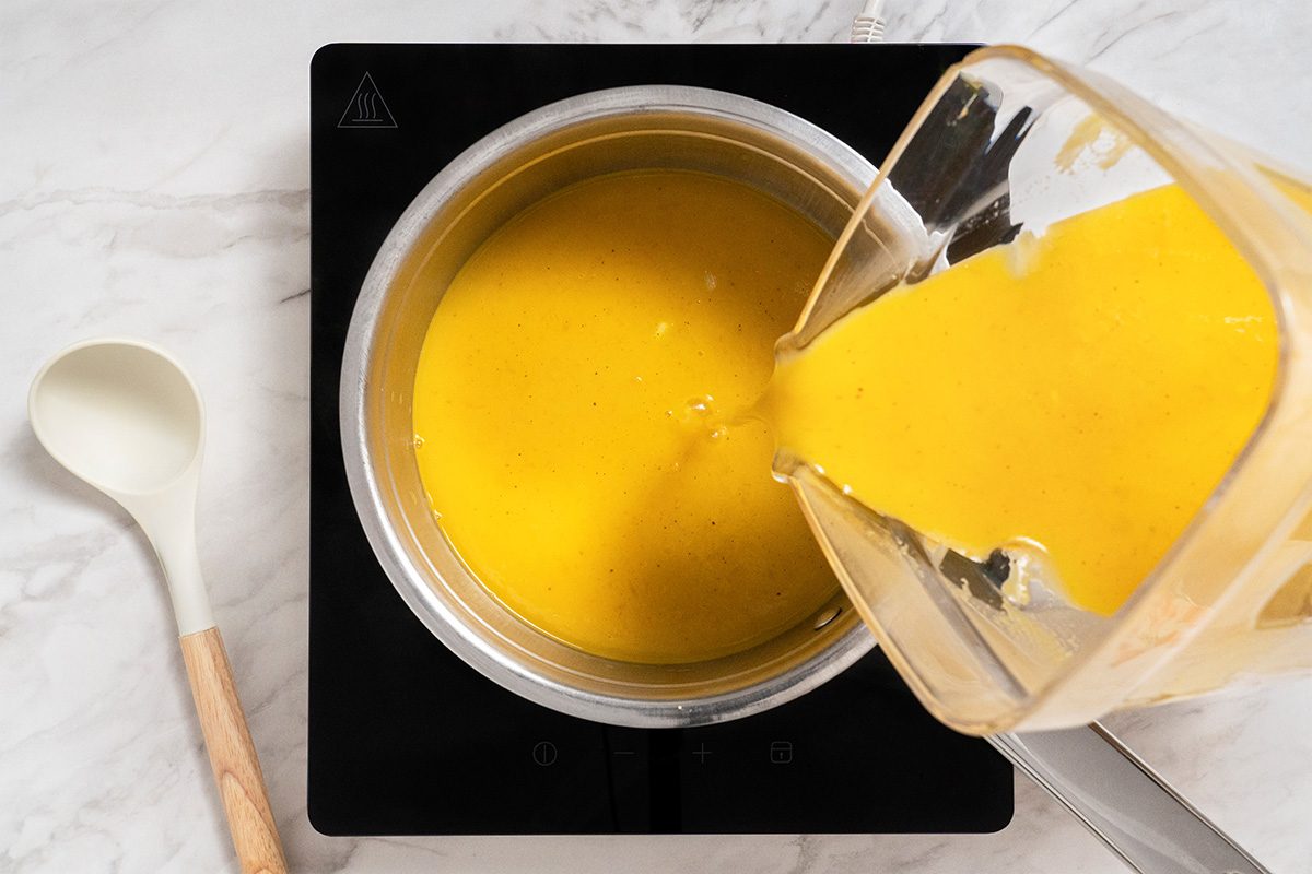 A blender is pouring yellow soup or sauce into a saucepan on an electric stovetop. A white spoon with a wooden handle rests on the countertop nearby.