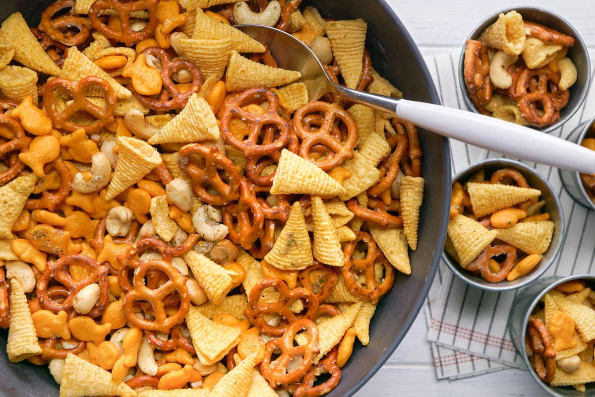 A bowl of snack mix; including pretzels; cheese crackers; corn chips; and nuts; sits with a spoon inside; surrounded by small metal cups of the mix on a white wooden surface
