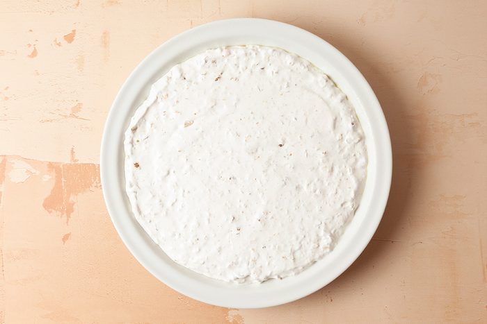 A white round dish filled with an even layer of creamy white dip, placed on a light beige surface.