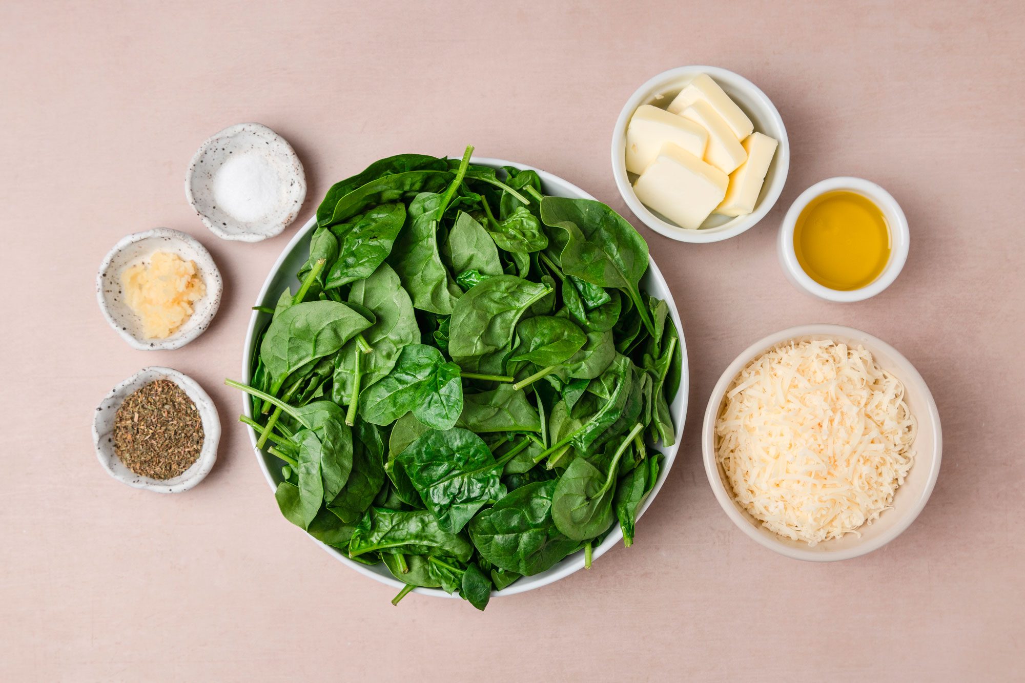 A top shot of a bowl of vibrant spinach is surrounded by small bowls containing butter; olive oil; shredded cheese; dried herbs; minced garlic; and salt on a light brown surface, creating a fresh and inviting meal preparation scene