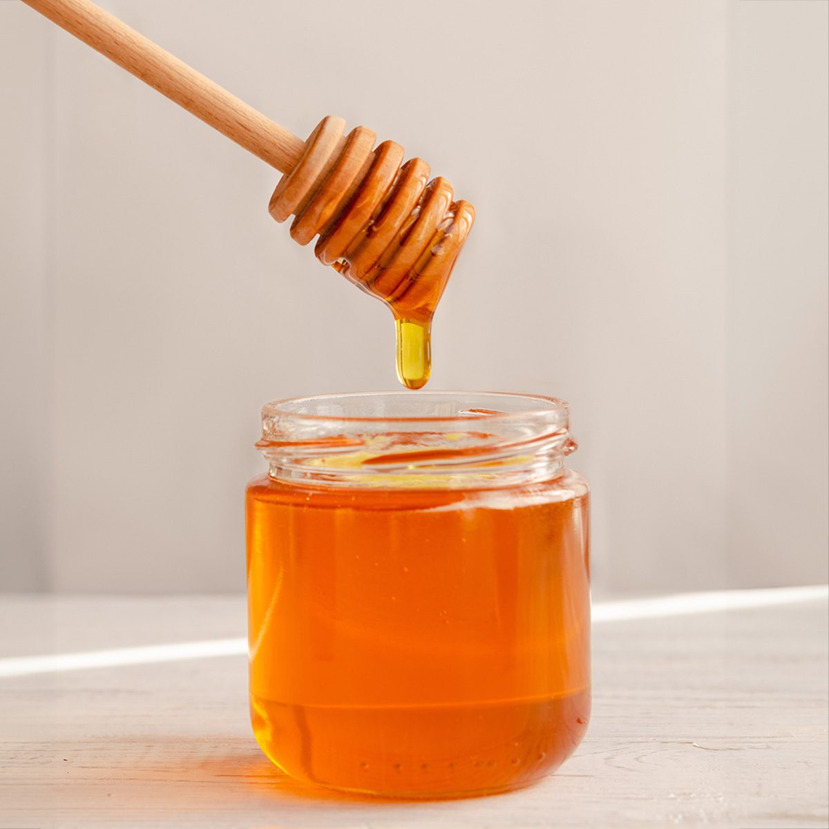 A viscous stream of sweet amber flower honey flows from a wooden honey spoon into a glass jar