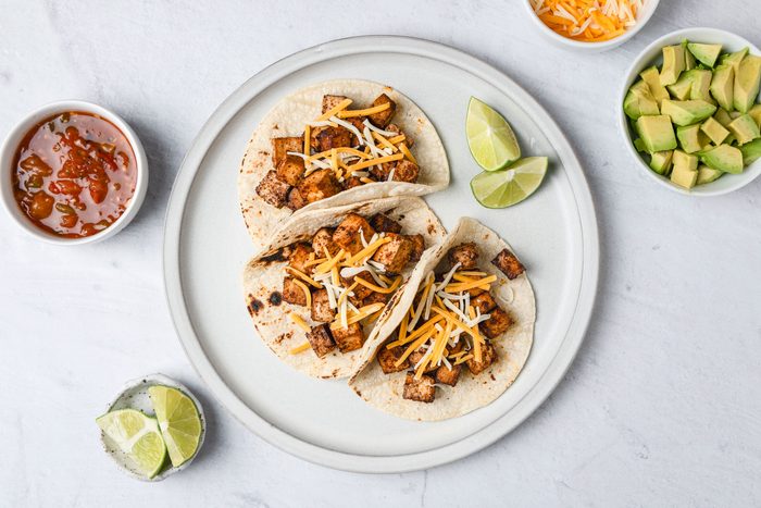 A top shot of a Three tacos filled with grilled tofu and shredded cheese sit on a plate, garnished with lime wedges; the background shows bowls of salsa; shredded cheese; diced avocado; and a small dish of lime