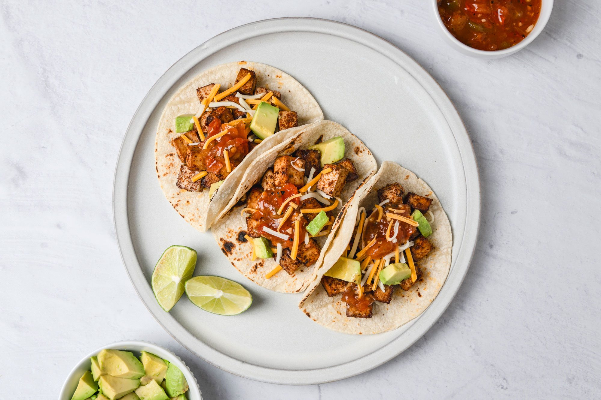 A top shot of a Three soft tacos filled with grilled chicken; cheese; avocado; and salsa are presented on a white plate with lime wedges; accompanied by bowls of salsa and diced avocado on a white background