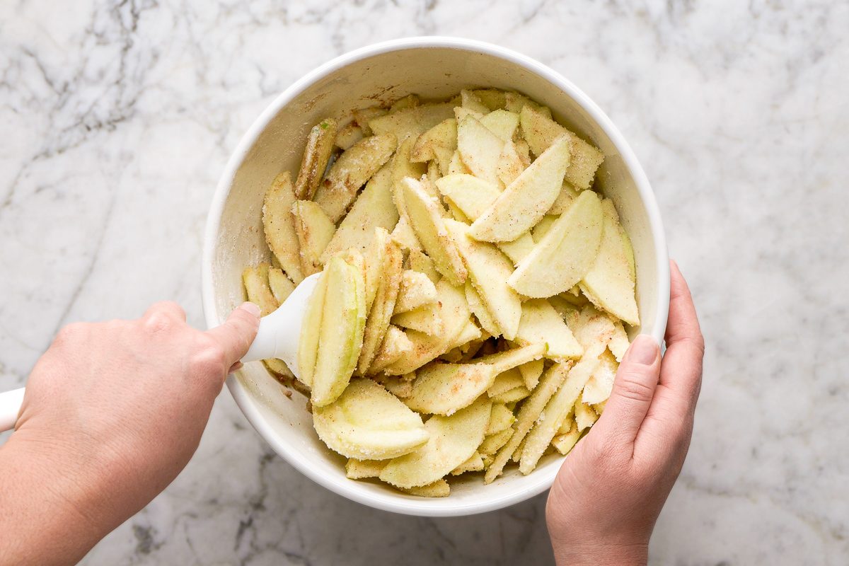 sliced apples, granulated sugar, flour, cinnamon and nutmeg being mixed with melted butter in a bowl