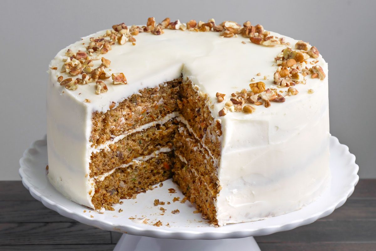 3/4 angle view of Zucchini Carrot Cake on a cake tray; A large slice has been removed; arranged on a dark brown wooden surface;
