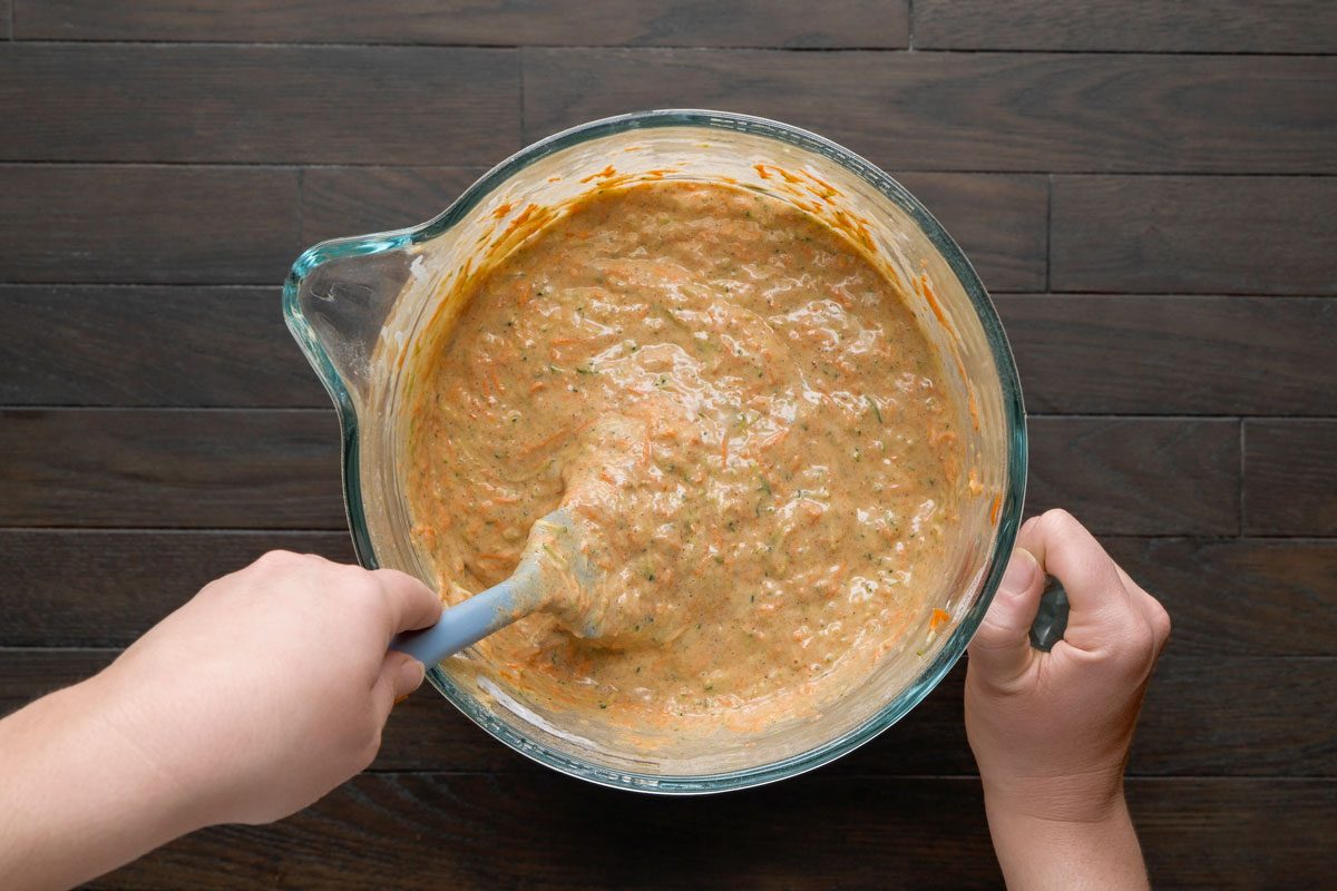 Overhead shot of a large glass jar; Stir in carrots; zucchini and nuts with spatula; all set on a dark brown wooden surface;