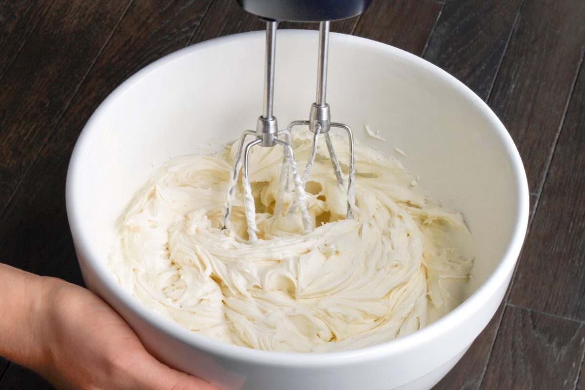 Close shot of a large bowl; For frosting beat cream cheese and butter in a large bowl until smooth; Add sugar and vanilla; Continue beating until sugar is dissolved; hand blender; all set on adark brown wooden surface;