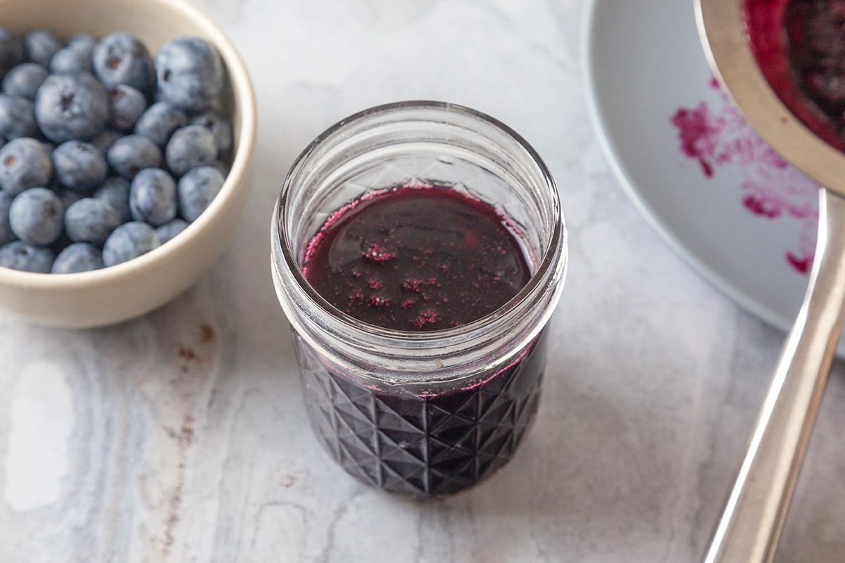 Overhead shot of a glass jar filled with dark blueberry sauce sits on a light surface; next to a bowl of fresh blueberries and a plate with sauce residue