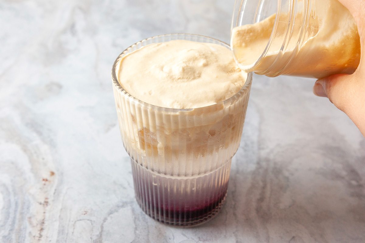 Close-up shot of cream being poured over a dark iced beverage in a ribbed glass, set on a light marbled surface
