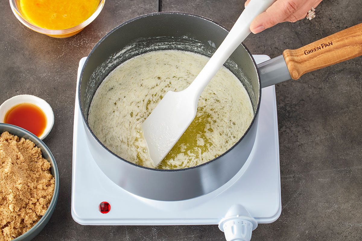 Overhead shot of a hand stirring melted butter with a white spatula in a saucepan on a hot plate; with bowls of brown sugar; a yellow liquid; and vanilla extract nearby;
