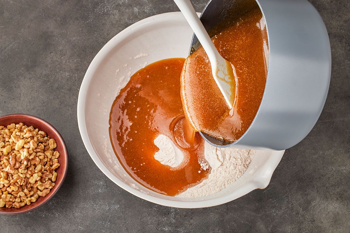 Overhead shot of caramel sauce being poured from a saucepan into a mixing bowl with flour; with a small bowl of chopped walnuts nearby on a gray countertop;