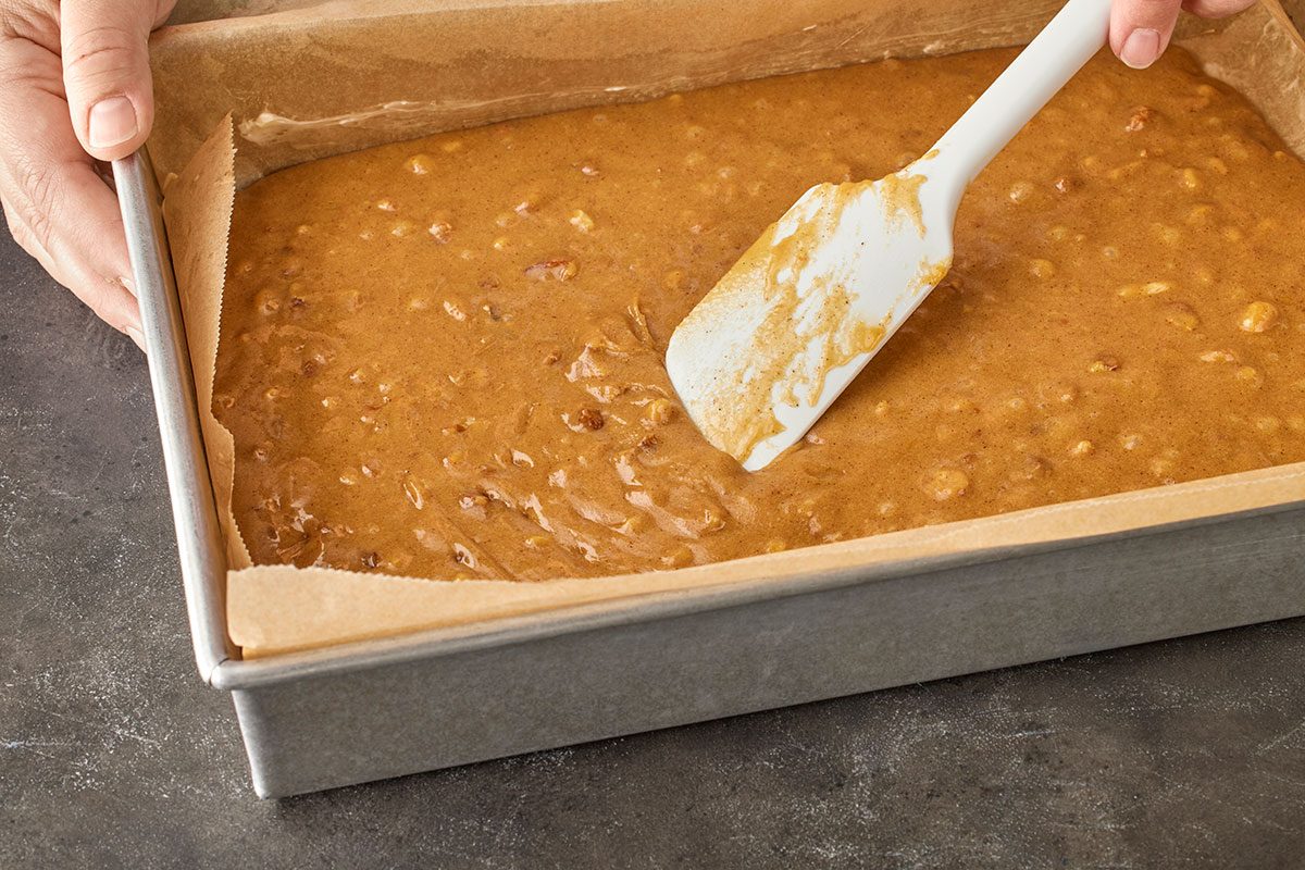 3/4 angle view shot of a person spreading brown batter in a parchment-lined baking pan with a white spatula on a dark countertop;