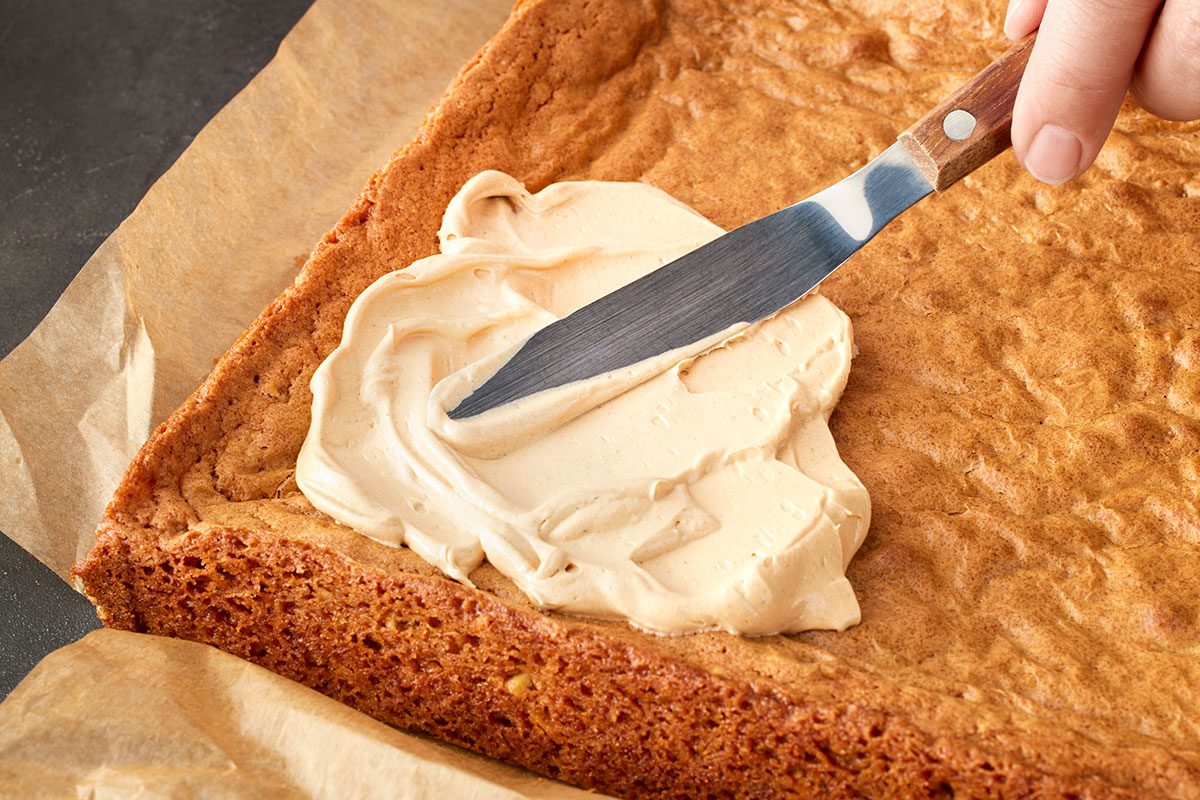 Close-up shot of a hand using a metal spatula to spread light brown frosting over a large rectangular sheet cake on parchment paper;