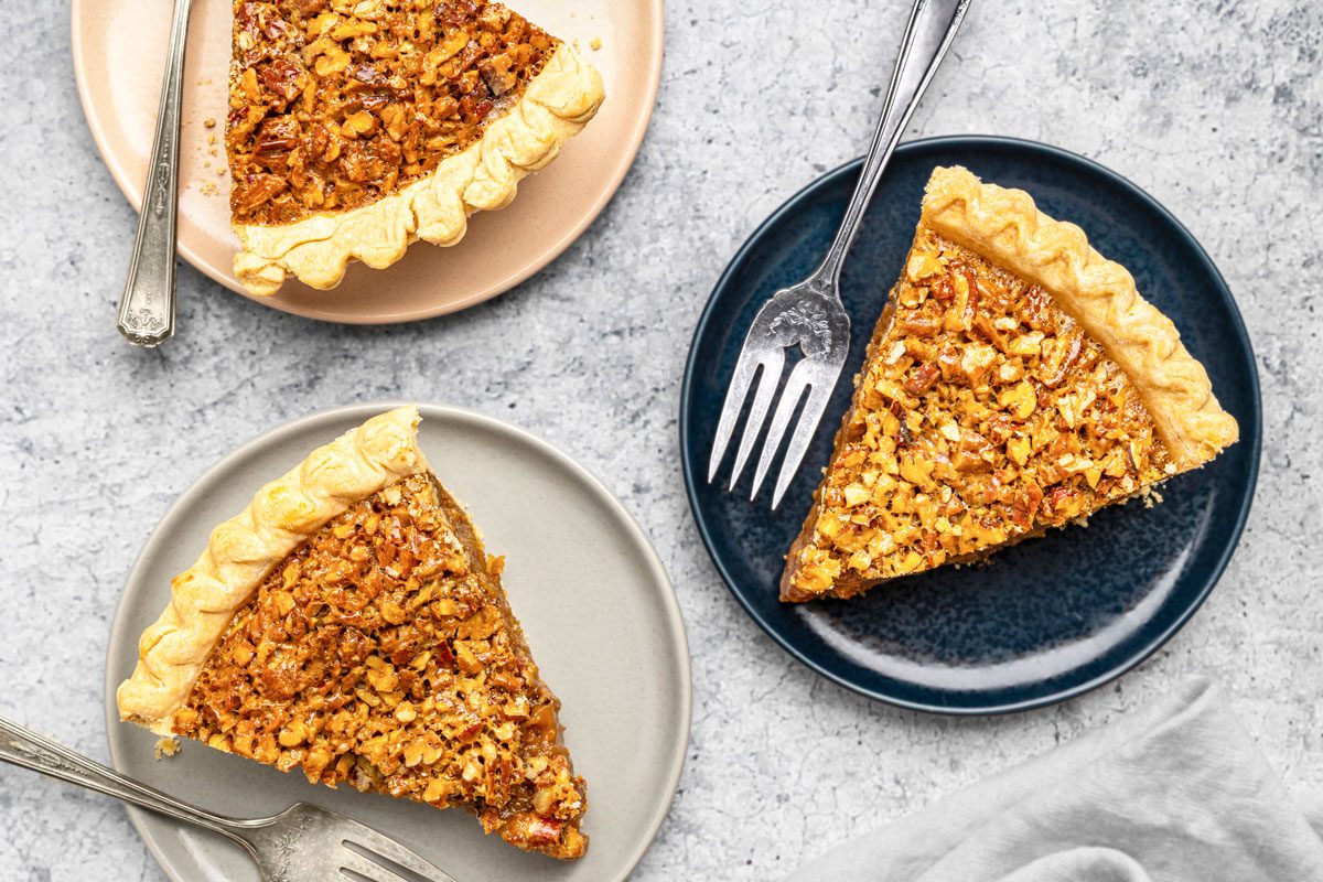 overhead shot of Three slices of pecan pie on separate plates with forks sit on a light textured surface, The pies have a golden, flaky crust and a nutty filling, Two plates are beige and one is dark blue