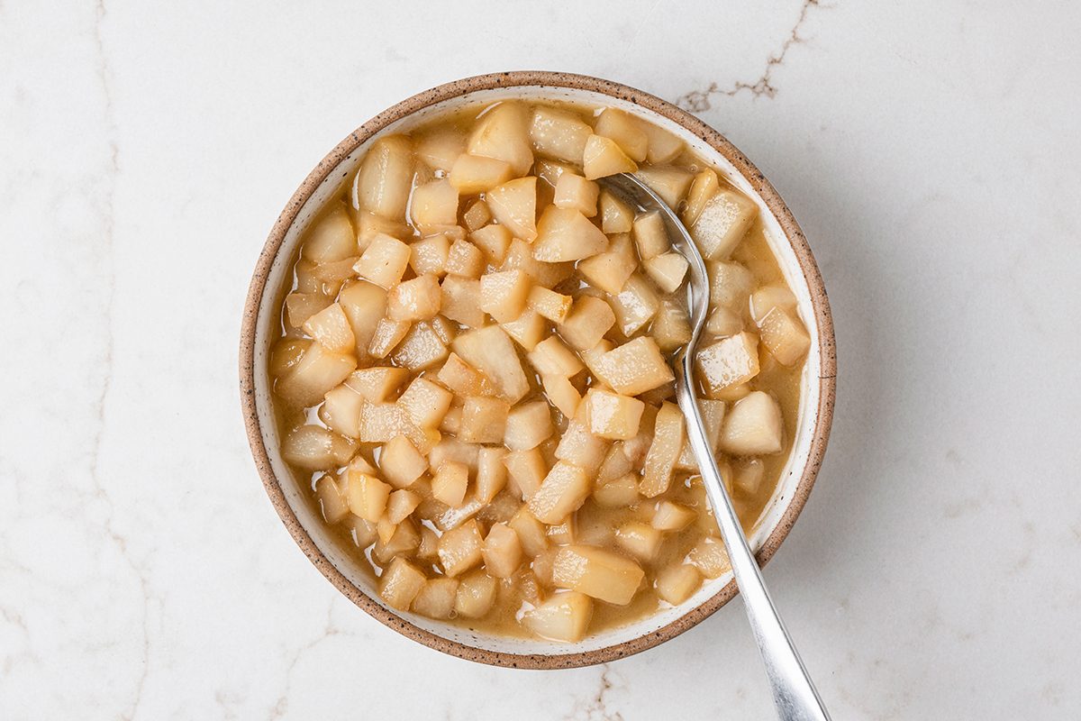 A bowl of diced cooked apples in syrup, with a spoon resting inside, placed on a light marble surface.