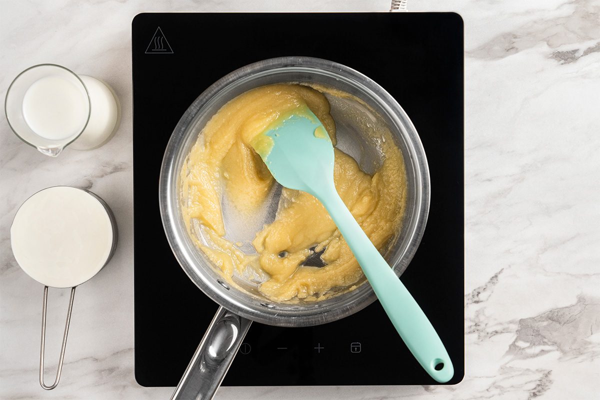 A saucepan on a stovetop contains a light brown roux being stirred with a light blue spatula. Next to the stove, there is a measuring cup of milk and a measuring cup of flour on a marble countertop.