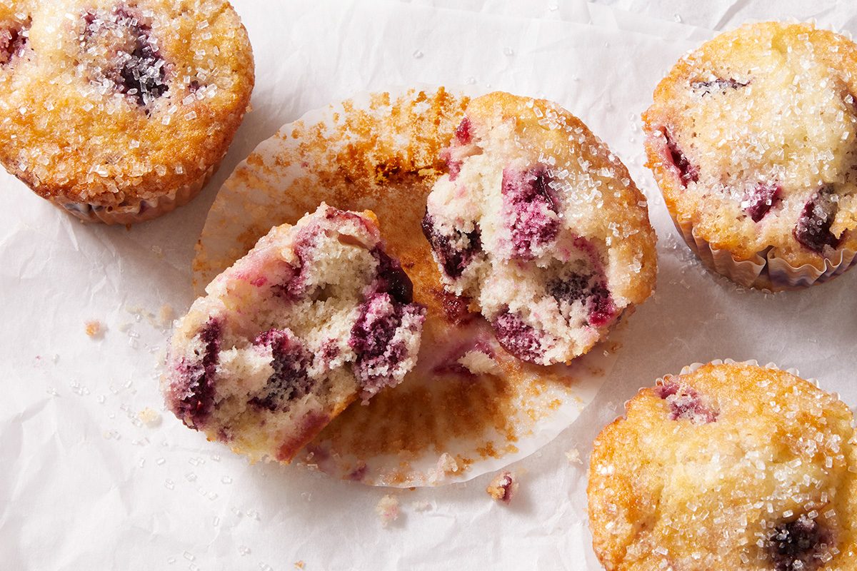Four golden-brown berry muffins are on parchment paper; one is cut in half, revealing a moist interior with purple berries. Sugar crystals are sprinkled on top, and there are crumbs on the paper.
