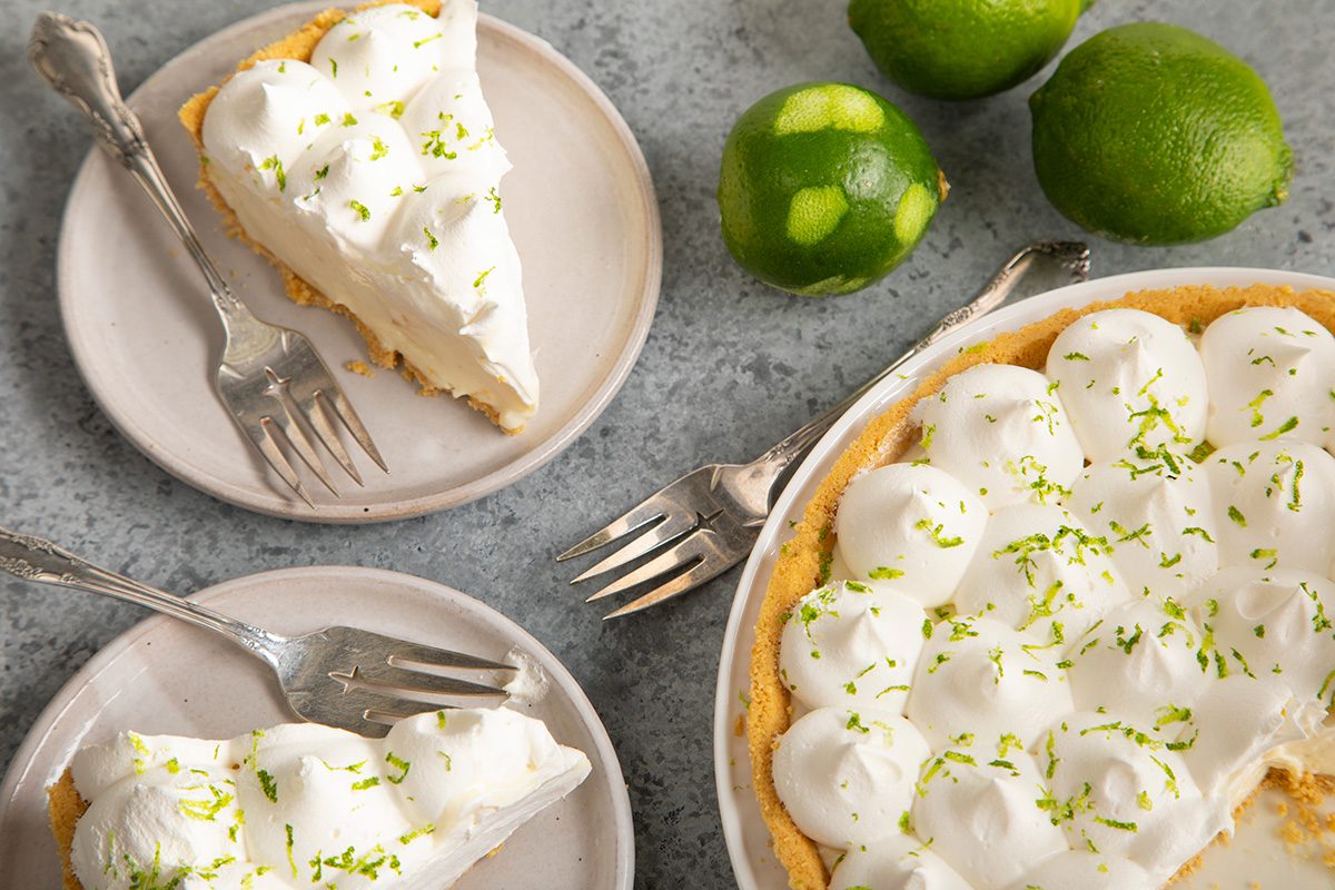 A key lime pie topped with whipped cream and lime zest is served on two plates with forks. Whole limes are nearby on a gray surface.
