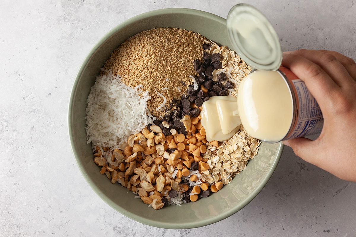 A hand pours sweetened condensed milk from a can into a bowl containing oats, shredded coconut, chocolate chips, chopped nuts, and sesame seeds on a light surface.