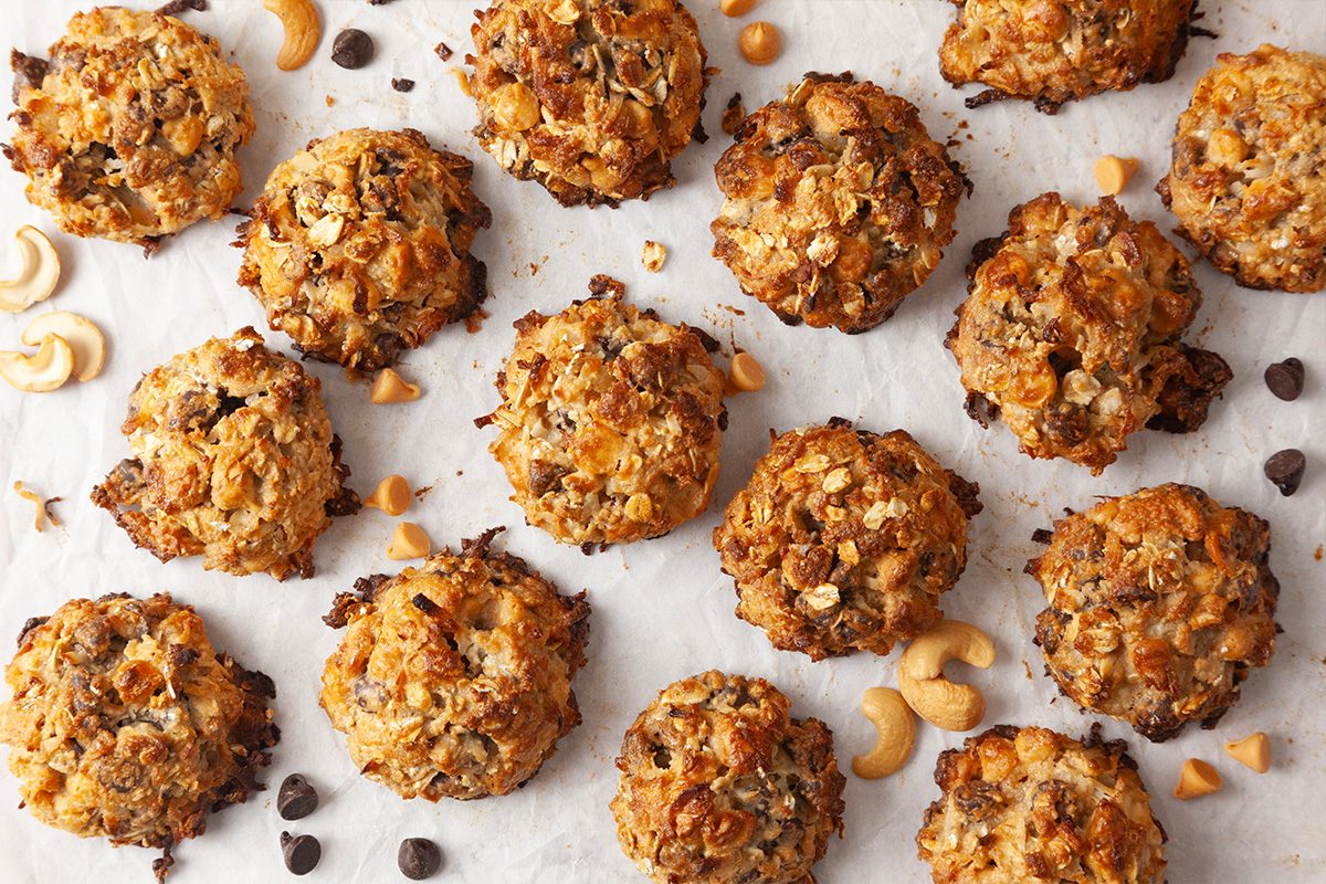 A batch of oatmeal cookies with chocolate chips and cashews is arranged on a parchment-lined surface, with scattered chocolate chips and cashew pieces around them.