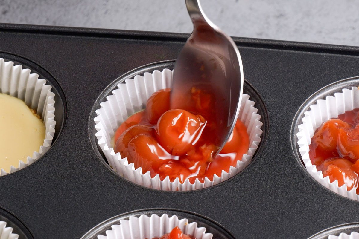 closeup shot of spoon is adding cherry pie filling into a paper cupcake liner placed in a muffin tin, preparing for baking