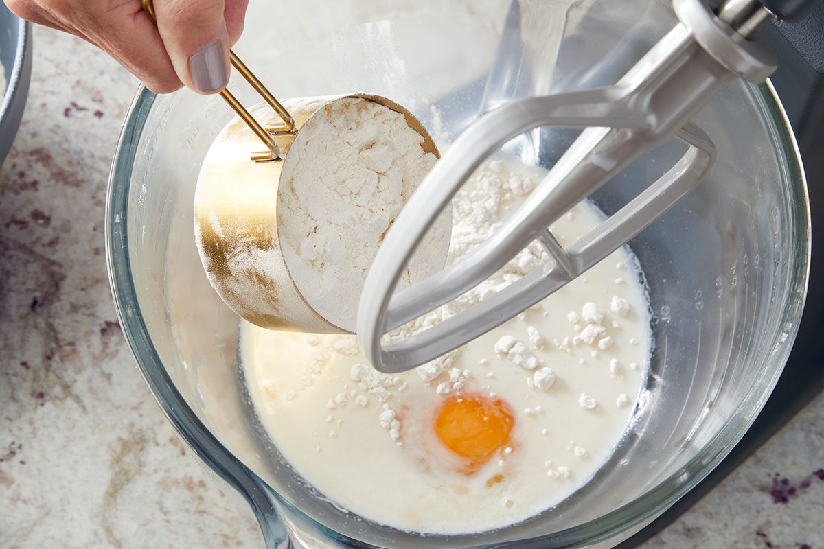 Overhead shot of a hand pouring flour from a measuring cup into a stand mixer bowl containing milk and a cracked egg; preparing ingredients for baking.