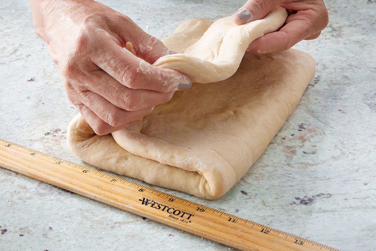 Close shot of a person folding a sheet of dough on a floured surface beside a wooden ruler.