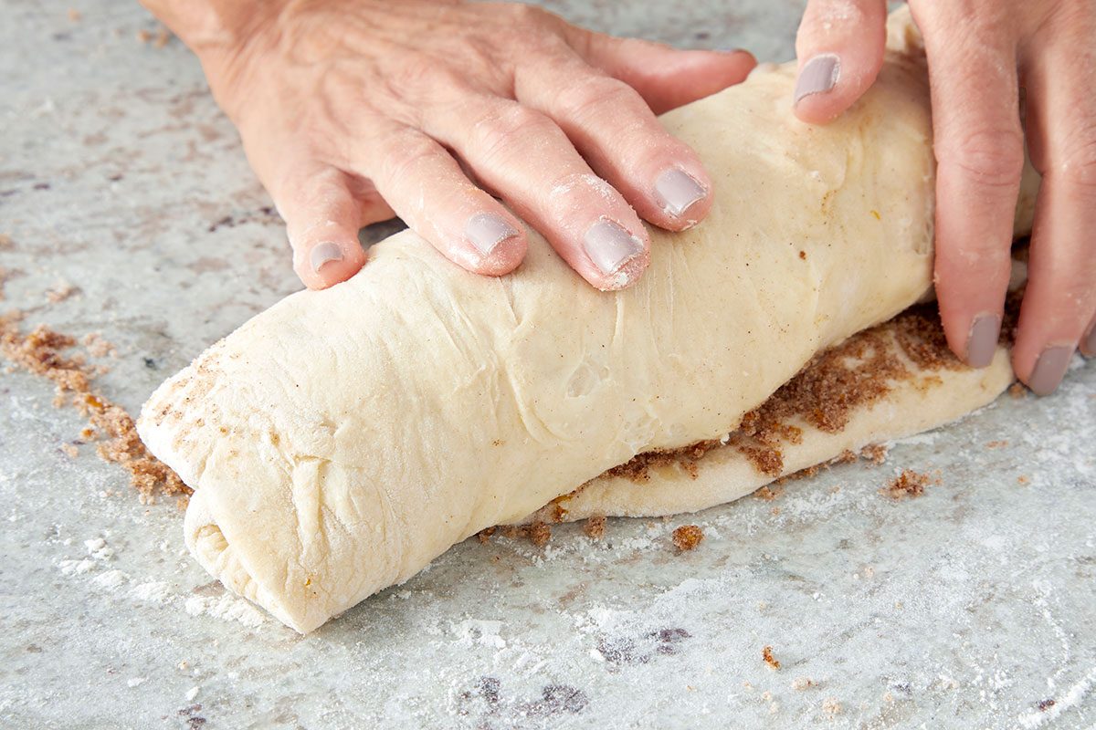 Close-up shot of hands with painted nails rolling up dough filled with cinnamon sugar on a floured surface; preparing cinnamon rolls;
