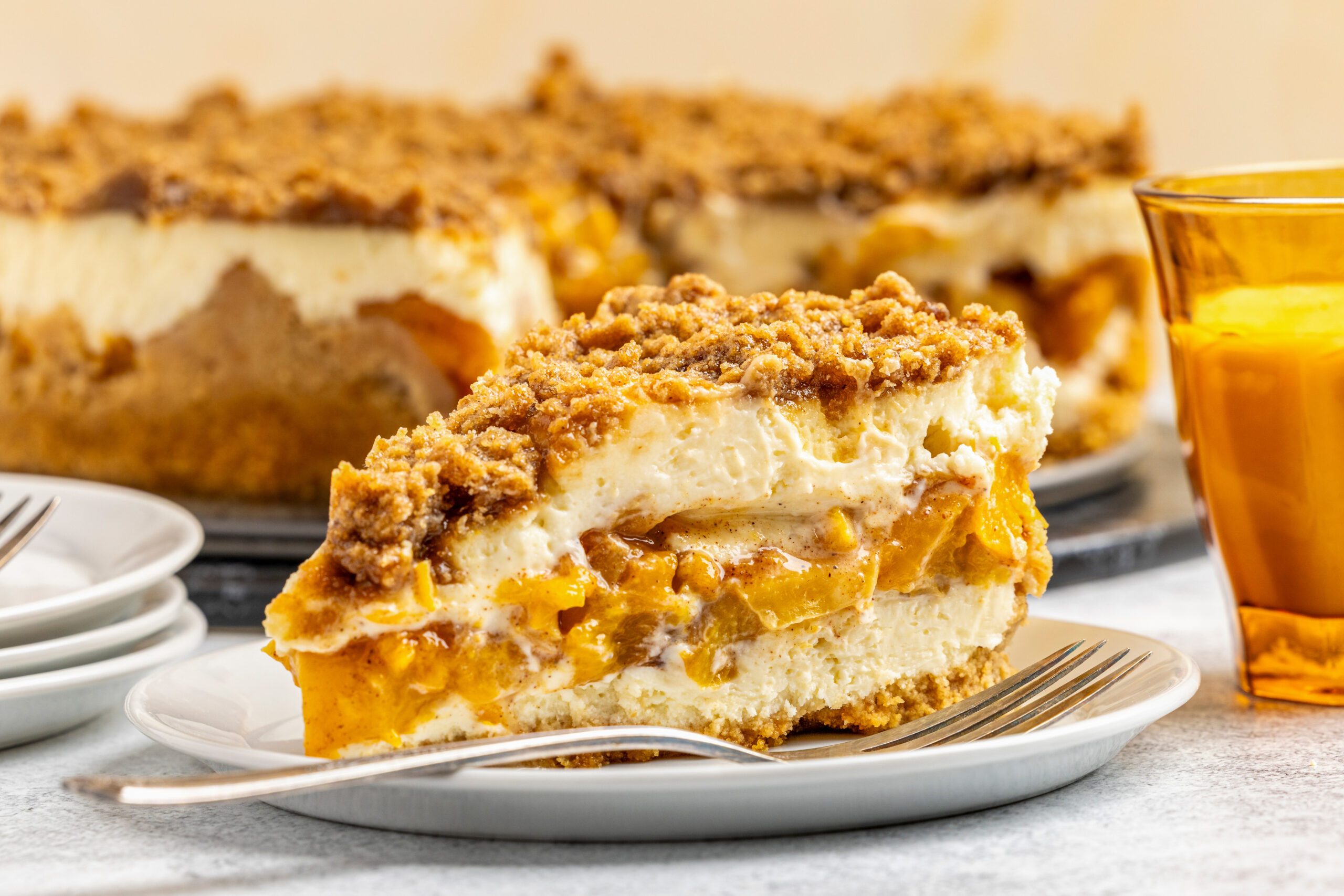 A slice of crumb-topped cheesecake with layers of peaches and cream sits on a white plate with a fork. More cheesecake and a glass of orange juice are in the background.